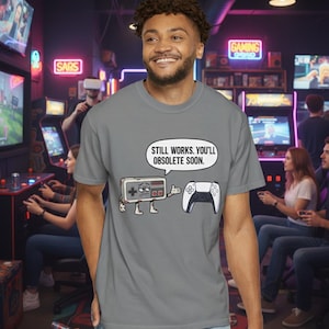 Peut inclure: T-shirt gris avec un dessin animé d'une manette de jeu rétro et d'une manette moderne. Le texte sur le t-shirt se lit "Still works. You'll obsolete soon."
