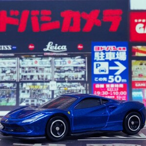 May include: A blue toy car model of a Ferrari 488 Pista. The car is on a black and white surface with Japanese text in the background.