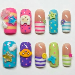 May include: A collection of ten artificial nails, each with a unique design. The nails showcase vibrant colours including pink, blue, and green, adorned with polka dots, stripes, and 3D decorations like stars, clouds, and cartoon characters.