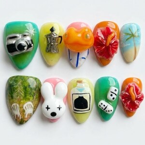May include: A collection of ten artificial nails, each with a unique design. Designs include a camera, a coffee pot, a frog, a hibiscus flower, a beach scene, a forest scene with chairs, a bunny, a perfume bottle, dice, and another hibiscus flower. The nails are in various colours.