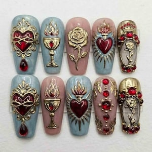 May include: A set of ten artificial nails with ornate designs. The nails feature gold-colored embellishments, including hearts, roses, and chalices, with red and blue accents. The nail colors vary from light blue to pink.