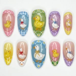 May include: A set of ten artificial nails with a duck theme. Each nail features a different design, including yellow ducklings and white ducks with bows, set against pastel backgrounds of pink, blue, green, and purple. The nails are oval-shaped.