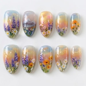 May include: A set of ten almond-shaped press-on nails featuring a hand-painted landscape design. The artwork displays a soft sunset sky background with various floral motifs including purple lupines, white daisies, and orange wildflowers.