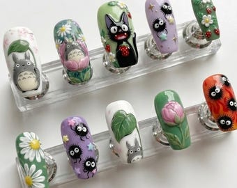 Könnte beinhalten: Ein Set aus zehn künstlichen Fingernägeln mit detaillierten, handgemalten Designs. Die Nägel zeigen verschiedene Charaktere und Motive, darunter Totoro, Rußgeister und florale Elemente, in einer Vielzahl von Farben. Jeder Nagel ist auf einem transparenten Präsentationsständer montiert.
