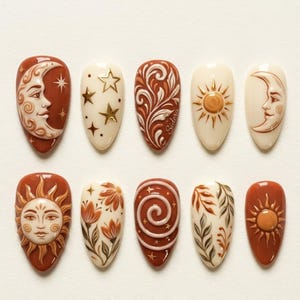 May include: A set of ten almond-shaped artificial nails with celestial designs. The nails feature a color palette of terracotta, cream, and gold. Designs include suns, moons, stars, and floral patterns, with intricate details and a glossy finish.