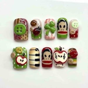 May include: A set of ten artificial nails with various designs. The designs include a green button, a ladybug, a four-leaf clover, a cartoon face, a red apple, and piano keys. The nails are decorated with polka dots, stripes, and other patterns.