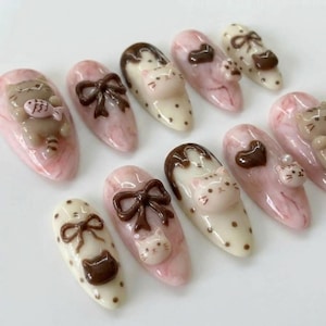 May include: A collection of ten artificial nails with various designs. The nails feature a pink and white marbled base, adorned with 3D cat and bow designs in brown and white. Some nails have a polka dot pattern.