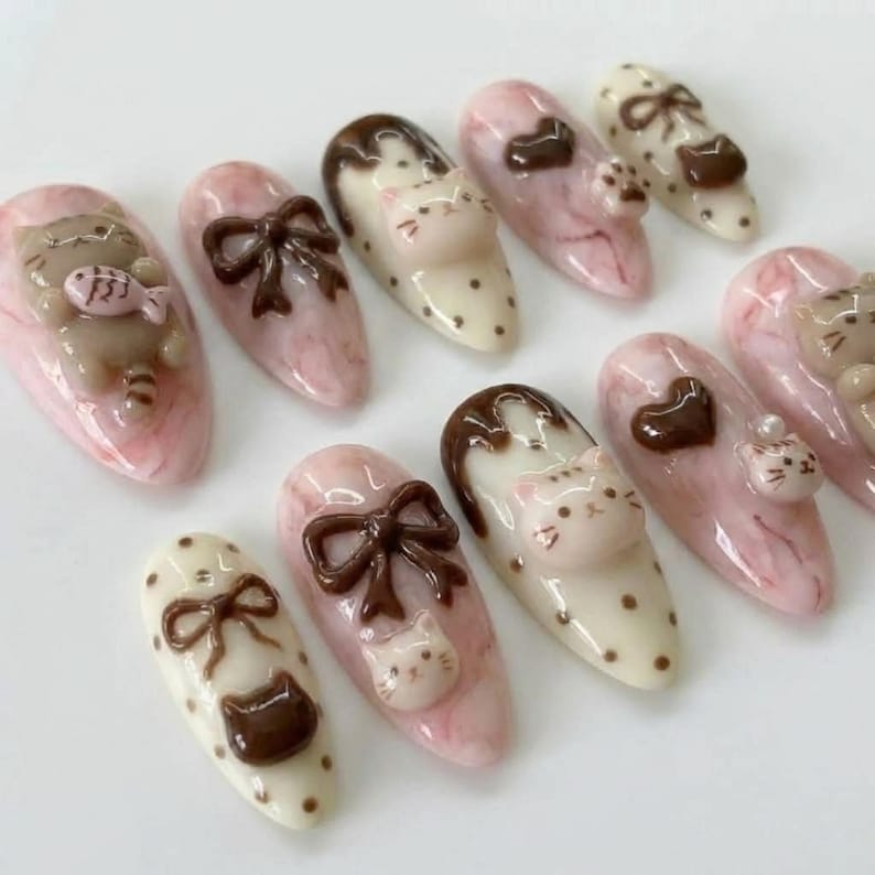 May include: A collection of ten artificial nails with various designs. The nails feature a pink and white marbled base, adorned with 3D cat and bow designs in brown and white. Some nails have a polka dot pattern.