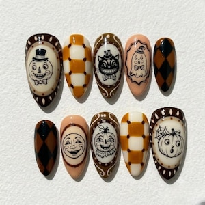 May include: A set of ten almond-shaped press-on nails featuring Halloween-themed designs. The collection includes illustrations of a jack-o'-lantern, a cat, a ghost, a moon face, and a pumpkin, alongside brown and cream checkered and diamond patterns.