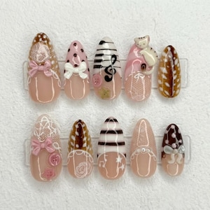 May include: A set of ten almond-shaped press-on nails featuring various designs including white lace patterns, pink bows, brown and white stripes, a small cat figure, musical notes, stars, and circular buttons in shades of pink, brown, and white.