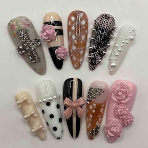 May include: A collection of ten artificial nails with various designs. The nails feature embellishments like silver studs, pink roses, pearls, bows, and black and white patterns. Some nails have a cross design, while others have a spiked pattern.