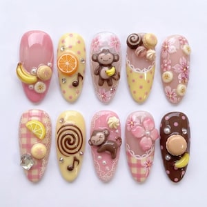 May include: A collection of ten artificial nails with diverse designs. The nails showcase a range of colours, including pink, yellow, and brown, and are adorned with fruit, musical notes, monkeys, and floral details.