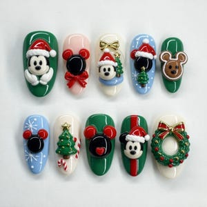 Könnte beinhalten: Ein Set aus zehn künstlichen Fingernägeln mit 3D-Weihnachtsdekorationen. Die Designs umfassen Mickey Mouse mit Weihnachtsmütze, Lebkuchenohren, einen grünen Weihnachtsbaum, eine rote Schleife und einen Kranz auf grünen, weißen und blauen Untergründen.