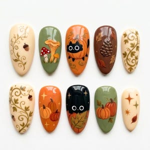 May include: A set of ten almond-shaped press-on nails featuring autumn-themed designs including a black cat in a pumpkin, mushrooms, pinecones, acorns, orange pumpkins, and gold star accents painted on cream, sage green, orange, and brown backgrounds.
