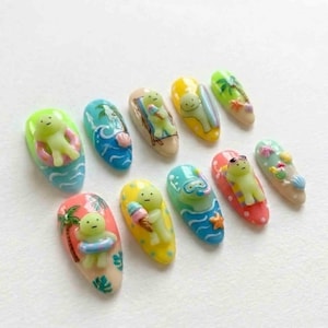 May include: A set of ten artificial nails, each with a unique summer-themed design. The nails feature miniature figures of a green character in various beach activities, including swimming, sunbathing, and enjoying ice cream. The nails are painted in bright colors.