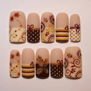 May include: A set of ten press-on nails featuring a matte nude base with autumn-themed designs including brown and gold maple leaves, white polka dots on dark brown, horizontal stripes, swirling patterns, and small red berry accents.
