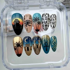 May include: A set of ten artificial nails with Egyptian-themed designs. The nails feature images of the Sphinx, Eye of Horus, palm trees, hieroglyphs, and other Egyptian symbols. The nails are in a clear plastic container.