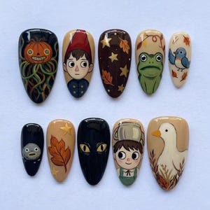 May include: A set of ten press-on nails featuring hand-painted designs including a pumpkin, a character in a red hat, gold stars, autumn leaves, a green frog, a blue bird, a dark rock-like figure, a black cat face, and a white duck.