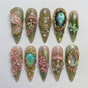 May include: A set of ten almond-shaped press-on nails featuring intricate 3D designs including pink butterfly wings, mushrooms, gold celestial patterns, iridescent gemstones, green ferns, pink roses, foxglove flowers, and clear crystals on a sage base.