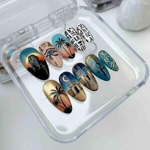 May include: A set of ten artificial nails with Egyptian-themed designs. The nails feature images of palm trees, pyramids, hieroglyphs, and the Eye of Horus. The color palette includes gold, black, blue, and peach.