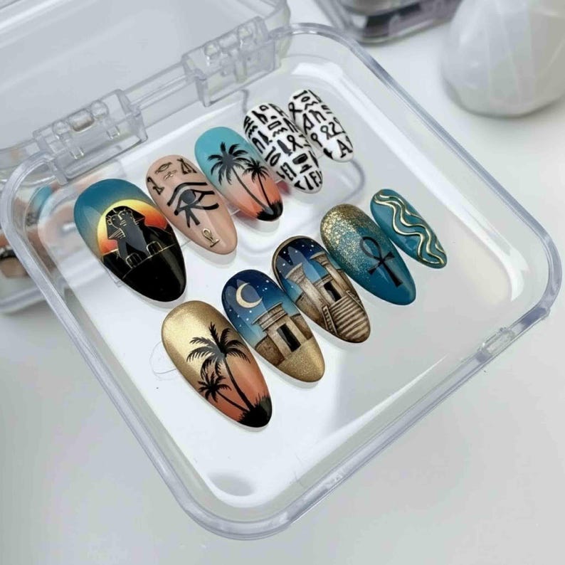 May include: A set of ten artificial nails with Egyptian-themed designs. The nails feature images of palm trees, pyramids, hieroglyphs, and the Eye of Horus. The color palette includes gold, black, blue, and peach.