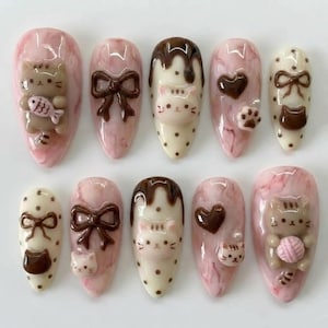 May include: A set of ten almond-shaped false nails with a pink marble effect. Each nail features 3D embellishments, including brown bows, cat faces, hearts, paw prints, and a cat holding a ball of yarn or a fish.