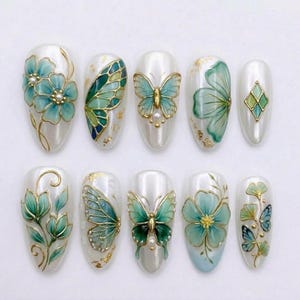 May include: A set of ten almond-shaped press-on nails with a white base, featuring hand-painted teal and green floral and butterfly designs, gold leaf accents, and small white pearl embellishments arranged in two horizontal rows of five.
