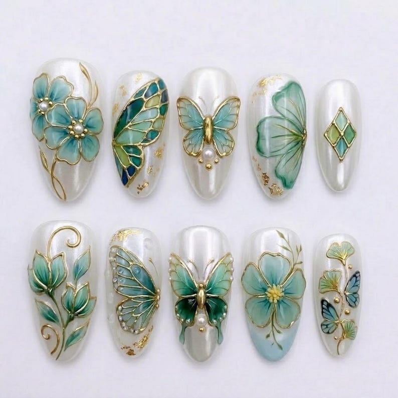 May include: A set of ten almond-shaped press-on nails with a white base, featuring hand-painted teal and green floral and butterfly designs, gold leaf accents, and small white pearl embellishments arranged in two horizontal rows of five.
