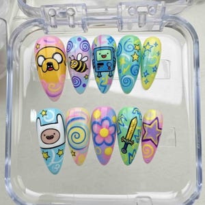 May include: A set of ten artificial nails with cartoon character designs. Each nail features a different design, including characters, stars, swirls, and flowers, in a variety of colors such as yellow, blue, pink, and green. The nails are displayed in a clear plastic case.