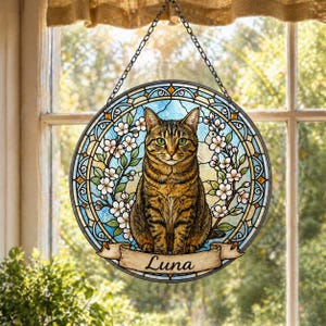 Op de afbeelding: Ronde glas-in-lood raamdecoratie met een tabby kat met groene ogen, omringd door witte bloemen en een blauwe achtergrond. De naam "Luna" staat op een banner. Het raam heeft een decoratieve rand en hangt aan een ketting.