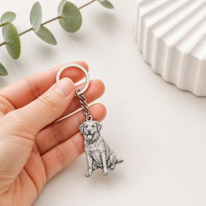 Puede incluir: Un llavero plateado con una ilustración detallada de un perro Labrador Retriever. El perro está representado en posición sentada con la lengua fuera. El llavero tiene un anillo circular y un eslabón de cadena.
