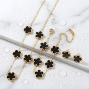 Può includere: Un set di gioielli color oro composto da collana, bracciale e orecchini. Ogni pezzo presenta un design a trifoglio con centri in smalto nero. Il set è esposto su una superficie bianca con goccioline d'acqua.