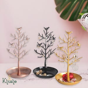 May include: Three decorative jewelry trees in rose gold, black, and gold. Each tree has a bird perched on top and branches for hanging jewelry. The trees have a round base to hold rings and other small items. The brand name "Khusha" is at the bottom.