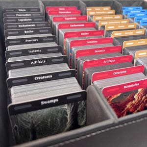 May include: A close-up of a gray storage box filled with stacks of trading cards. The cards are organized by category, with labels such as "Tokens," "Creatures," and "Mountains." The cards have red, black, and tan accents.
