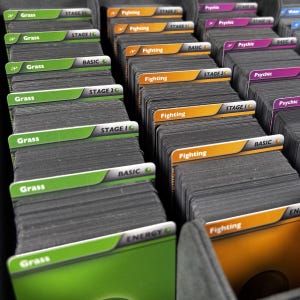 May include: Stacks of trading cards organized in a storage container. The cards are categorized by type, with labels like "Grass," "Fighting," and "Psychic." Each stack is labeled with "BASIC," "STAGE 10," or "STAGE 20."