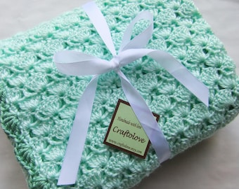 May include: A light green crocheted baby blanket with a white satin ribbon tied in a bow. The blanket has a scalloped edge and a tag that reads "Handmade with love, Craftolove, www.craftolove.etsy.com".