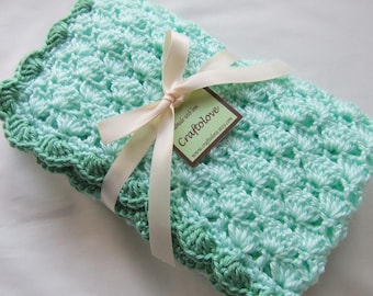 May include: A light green and dark green crocheted baby blanket with a white satin ribbon tied around it. The blanket has a scalloped edge and a tag that says "Craftolove" with the website address "www.craftolove.etsy.com".