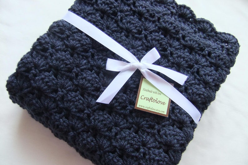 May include: A navy blue crocheted baby blanket with a white satin ribbon tied in a bow. The blanket is handmade and has a tag that says "Handmade with love, Craftlove, www.craftloveetsy.com".