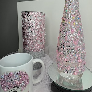 May include: A white ceramic mug with a pink and pearl design, a cylindrical container, and a decorative glass cone, all adorned with pink and silver embellishments. The items are arranged on a mirrored surface.