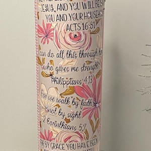 Puede incluir: Un portavelas decorativo cilíndrico con diseños florales y de escritura. El portavelas presenta flores rosas y blancas, con texto de versículos bíblicos. El portavelas está adornado con pequeñas cuentas rosas.