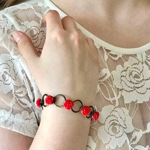 May include: A red rose bracelet with a silver chain and metal circles. The bracelet is on a person's wrist.