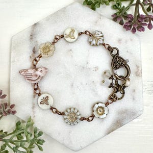 May include: A handcrafted bracelet featuring a variety of charms, including birds, bees, and flowers. The bracelet has a copper-colored chain and clasp. The charms are white with gold accents and pink details. The bracelet is approximately 7 inches long.