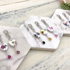 May include: A set of seven keychains featuring white porcelain heart charms with floral patterns. Each charm is attached to a silver chain with a crystal bead at the end. The charms are displayed on a white marble surface.