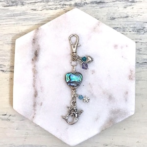 May include: A silver keychain with a heart-shaped abalone shell pendant. The keychain features a mermaid charm, a star charm, and various blue and purple beads. The abalone shell has iridescent green and blue hues.