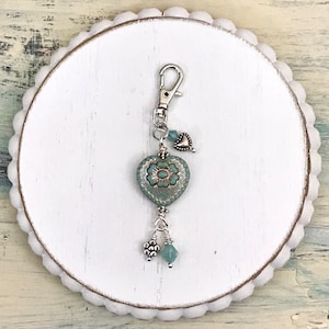 May include: A teal heart-shaped keychain with a floral design, silver accents, and dangling beads. The keychain features a lobster clasp for easy attachment. The heart pendant is adorned with a flower and small silver beads.
