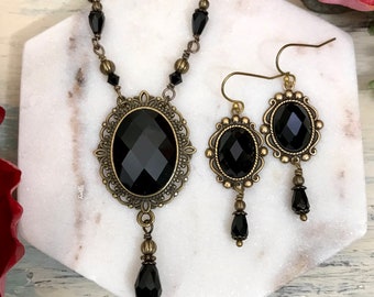 May include: A black and gold necklace and earrings set with a large oval black gemstone pendant and matching earrings. The necklace features a delicate chain with black beads and a dangling black gemstone drop.