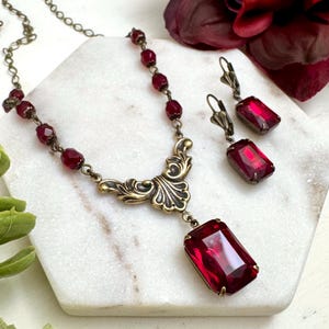 May include: A vintage-style jewelry set featuring a necklace and matching earrings. The necklace has a decorative brass filigree pendant with a large rectangular ruby-red crystal. The earrings also have ruby-red crystals. The set is displayed on a white surface.