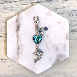 May include: A silver key chain with a blue and green abalone shell heart charm, a silver dolphin charm, and blue and purple crystal beads.
