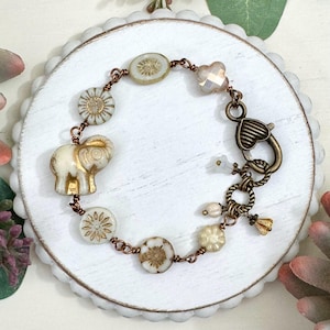 May include: A white and gold bracelet with a charm of an elephant, flowers, and a heart-shaped clasp. The bracelet is made with copper wire and has a vintage aesthetic.