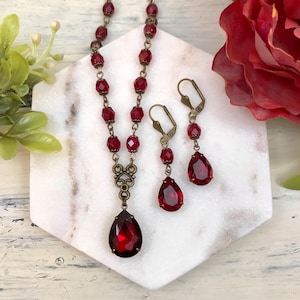 May include: A red crystal necklace and earrings set with a teardrop pendant and matching earrings. The necklace features a delicate chain with small red crystal beads and a vintage-inspired pendant. The earrings are simple and elegant, with a single teardrop crystal hanging from a small hook.